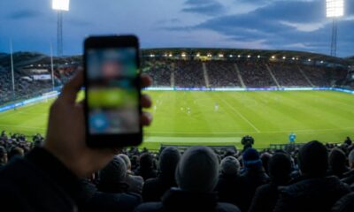 Supporter håller upp en mobiltelefon på läktaren under en fotbollsmatch i strålkastarljus