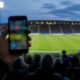 Supporter håller upp en mobiltelefon på läktaren under en fotbollsmatch i strålkastarljus