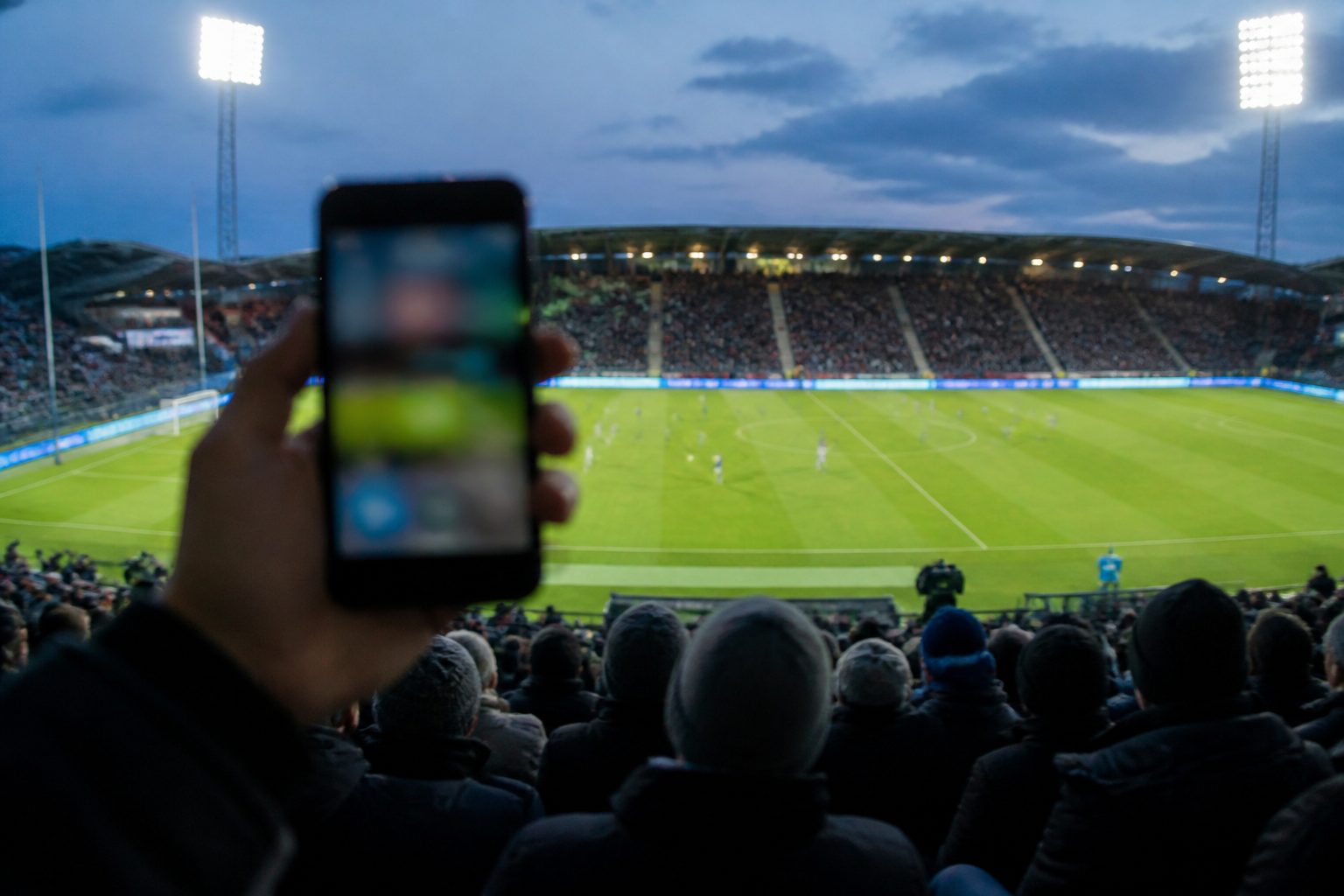 Supporter håller upp en mobiltelefon på läktaren under en fotbollsmatch i strålkastarljus